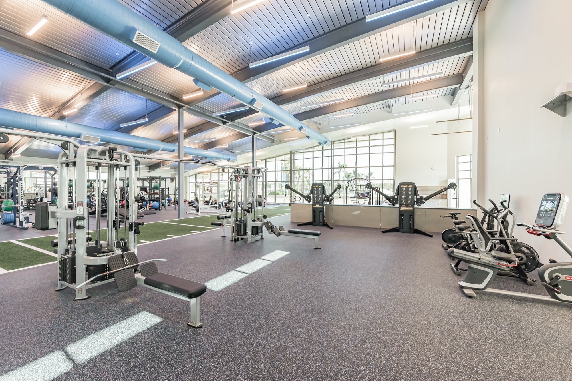 Open campus fitness floor with cable machines, benches and a turf training lane under an exposed ceiling