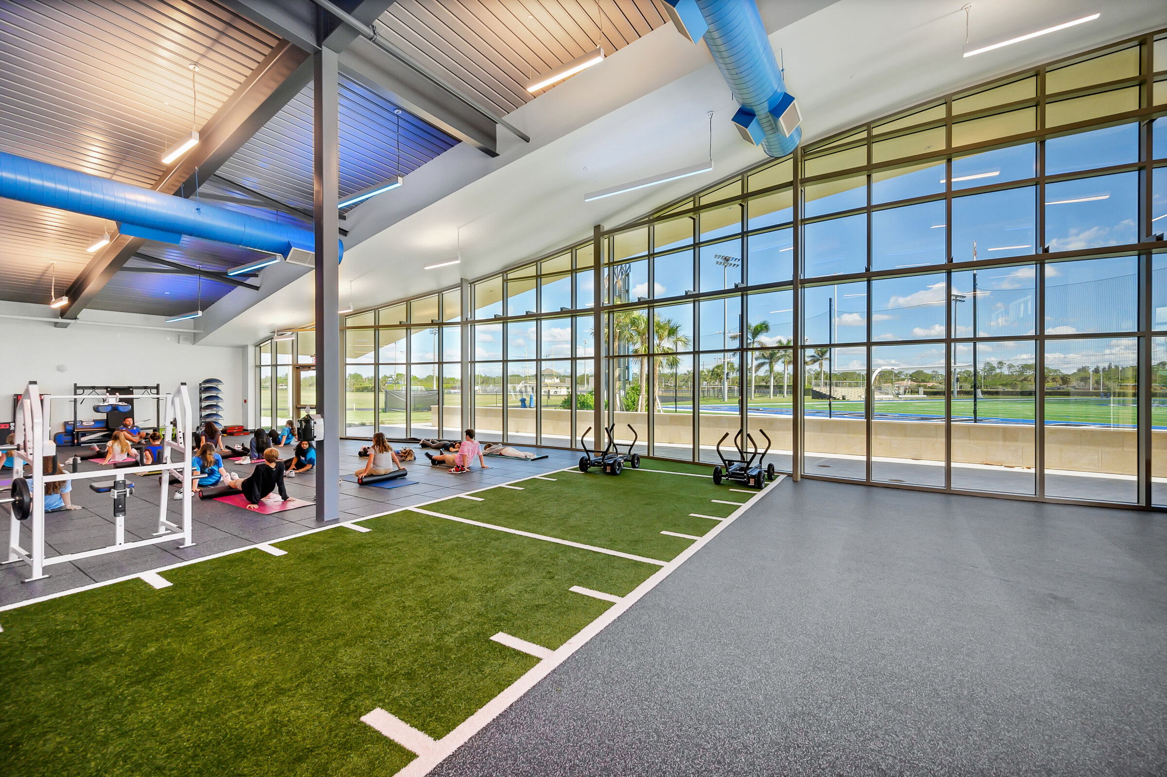 Sunlit fitness studio with turf training lanes and a stretching class beside floor-to-ceiling windows